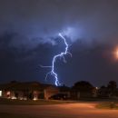 Lightning strikes above a suburban neighborhood's street at night. You can see several one story homes along the road.