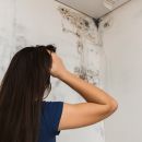 A woman puts her hand on her head as she gazes up at the corner of the wall in front of her. You can see mold growing from the ceiling and wall in front of her.