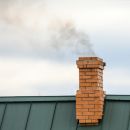 A closeup image of a green roof and brick chimney. You can see black smoke coming from the chimney.