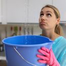 A woman looks up at the ceiling as she holds a blue bucket with pink gloves on. There is water leaking from the ceiling into the bucket