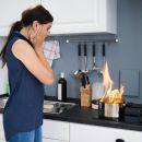 A woman stands in shock as a pot in her kitchen has flames eminating out of it