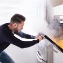 A man opens his oven door in shock as smoke billows out of it. You can see a small fire inside the oven.