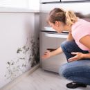 A woman looks shocked as she notices mold on the corner of the white wall in front of her