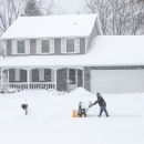 A closeup of a home during a snowstorm with a person using a snowblower on the driveway in front of it
