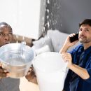 Two men look up at the ceiling in disgust while holding buckets. You can see water leaking down into the buckets