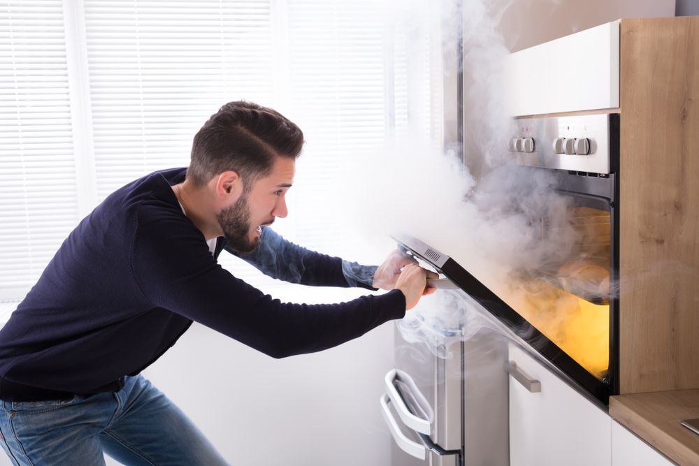 A man opens his oven door in shock as smoke billows out of it. You can see a small fire inside the oven.